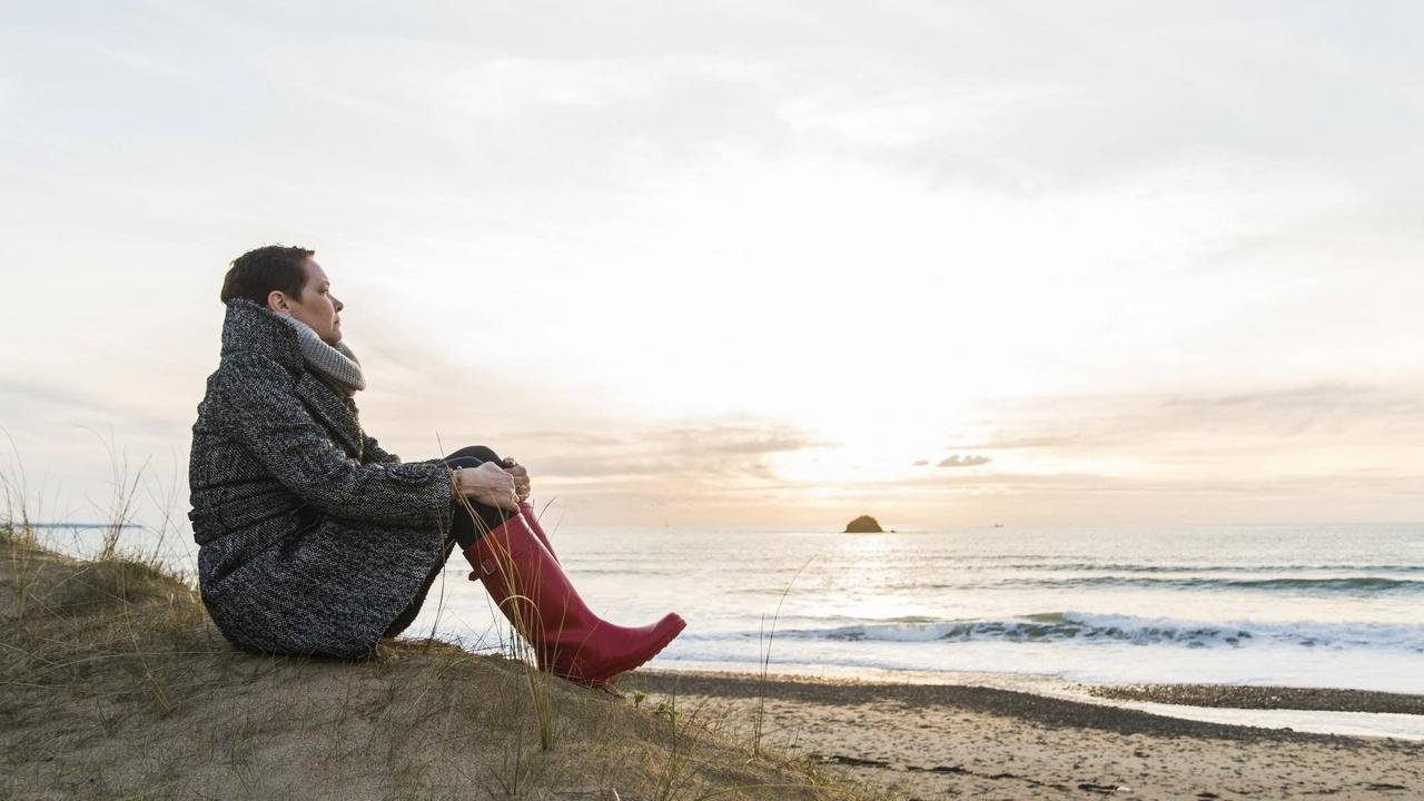 Eine Frau sitzt in Mantel und Gummistiefeln allein an einem Strand in Frankreich. Eine Frau sitzt in Mantel und Gummistiefeln allein an einem Strand in Frankreich.