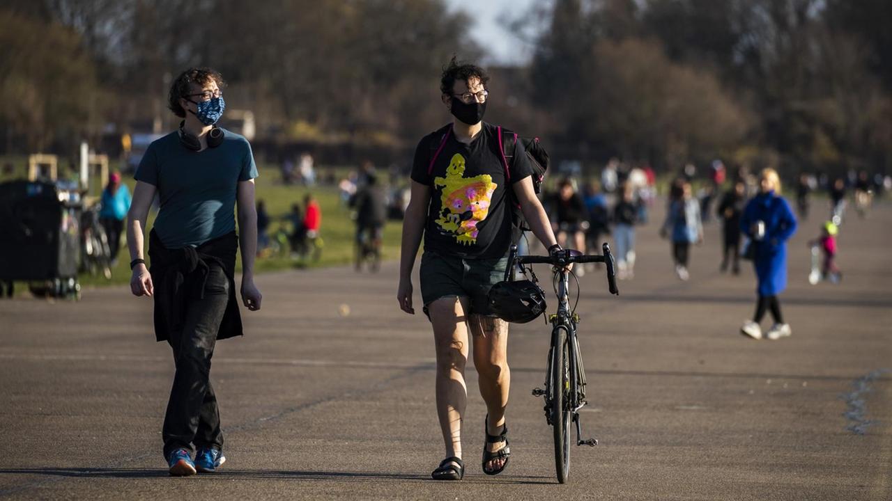 Zwei Spaziergänger mit Mundschutz, im Hintergrund mehrere Menschen bei schönem Wetter im Tempelhofer Feld Park in Berlin