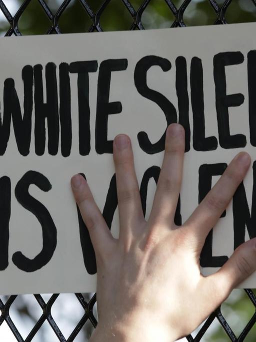 Eine Hand presst ein Schild mit der Aufschrift "White Silence is Violence" an einen Zaun.