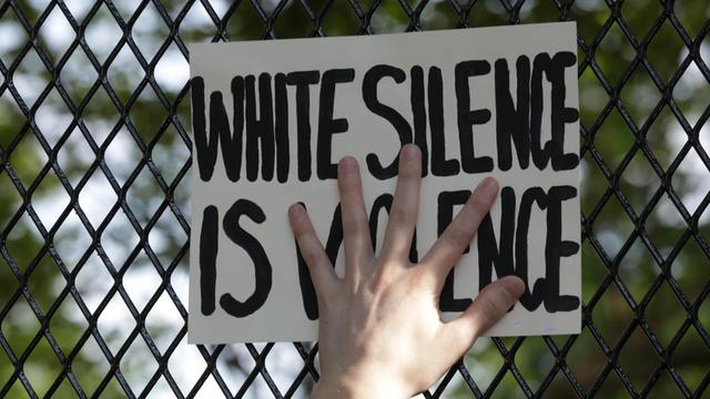 Eine Hand presst ein Schild mit der Aufschrift "White Silence is Violence" an einen Zaun.