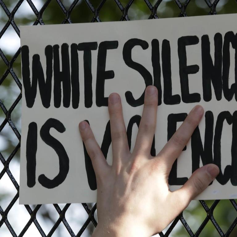Eine Hand presst ein Schild mit der Aufschrift "White Silence is Violence" an einen Zaun.