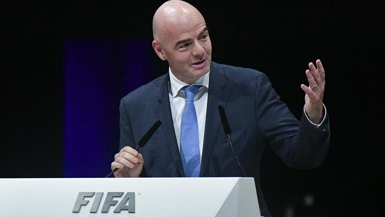 Infantino steht am FIFA-Redepult, lächelt, spricht und gestikuliert vor einem schwarzen Hintergrund.