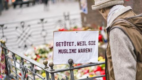 Eine Frau steht auf dem Marktplatz in Hanau vor dem Denkmal der Brüder Grimm. Vor dem Denkmal stehen Blumen, Kerzen und ein Plakat mit der Aufschrift «Getötet, weil sie Muslime waren».