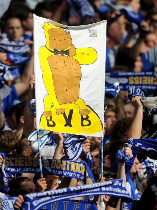 Schalke-Fans halten vor Spielbeginn ein Banner hoch, auf dem ein Affe in einem gelben Kostüm auf dem BVB-Vereinswappen sitzt.