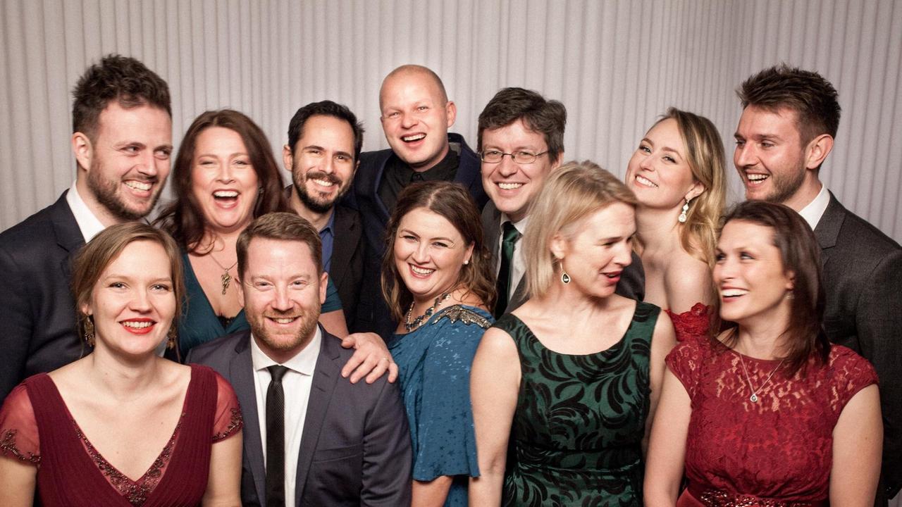 Die zwölf Ensembelmitglieder stehen in einer Gruppe und lachen beherzt in die Kamera. Die zwölf Ensembelmitglieder stehen in einer Gruppe und lachen beherzt in die Kamera.