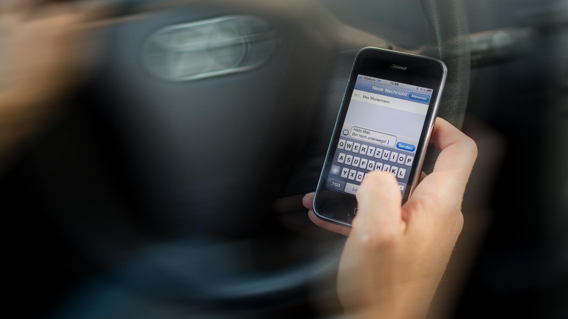 Eine Hand tippt vor einem Auto-Lenkrad in ein Handy. Eine Hand tippt vor einem Auto-Lenkrad in ein Handy.