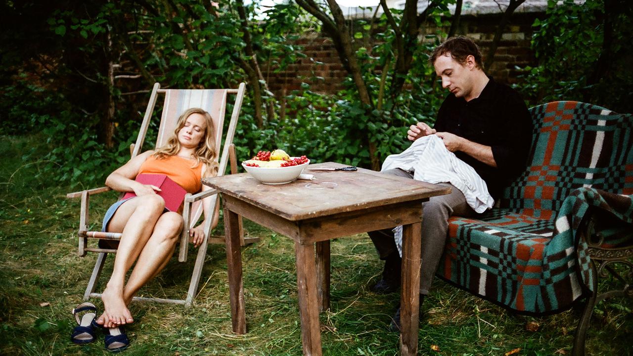 Adam (Florian Teichtmeister) versenkt sich in seine Nähkreationen, während Evelyn (Anne Kanis) in seinem Garten ein Mittagsschläfchen hält.