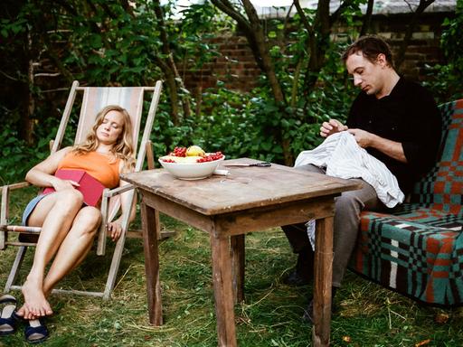 Adam (Florian Teichtmeister) versenkt sich in seine Nähkreationen, während Evelyn (Anne Kanis) in seinem Garten ein Mittagsschläfchen hält.