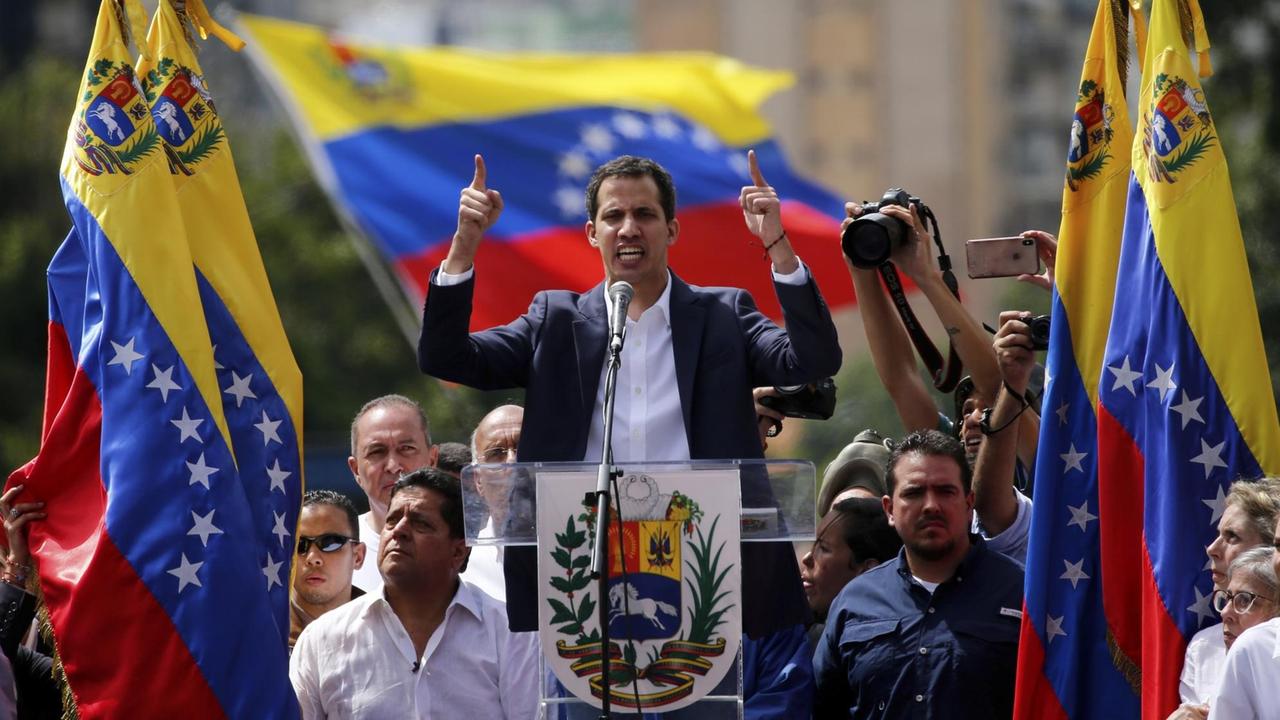Ein Mann spricht an einem Rednerpult, um ihn herum sind Flaggen des Landes Venezuela.
Ein Mann spricht an einem Rednerpult, um ihn herum sind Flaggen des Landes Venezuela.