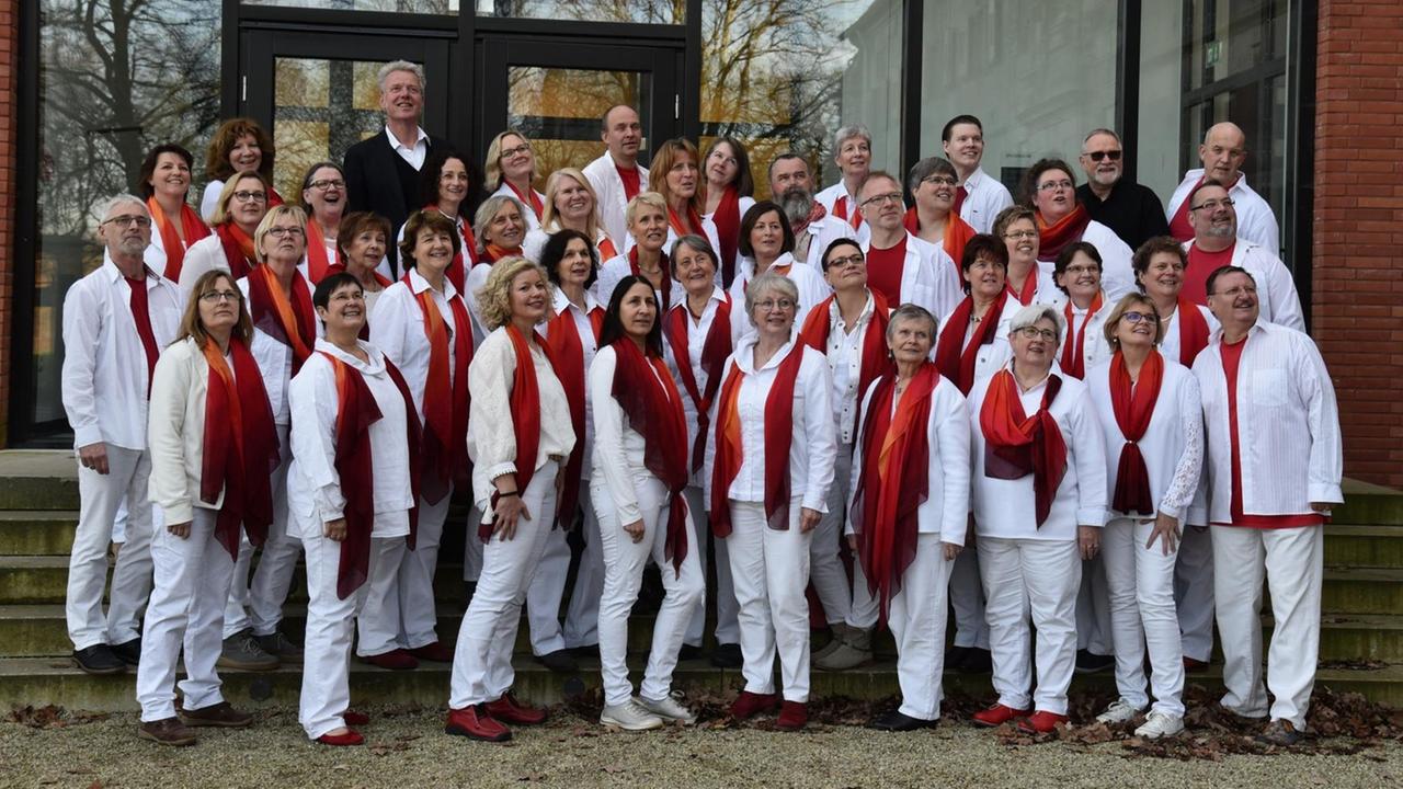 Gruppenbild des Chores vor einem Gebäude. Alle Sängerinnen und Sänger tragen weiße Kleidung und ein rotes T-Shirt. Gruppenbild des Chores vor einem Gebäude. Alle Sängerinnen und Sänger tragen weiße Kleidung und ein rotes T-Shirt.