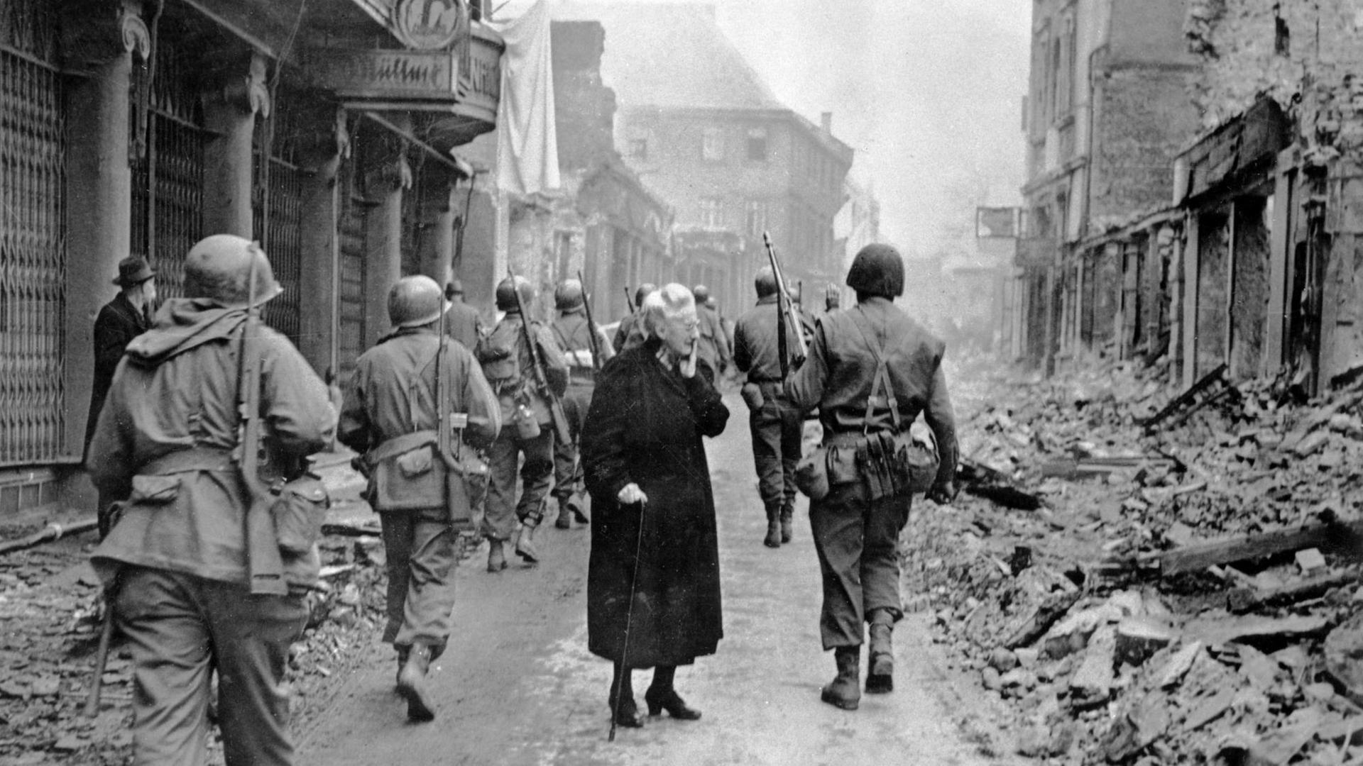 Fassungslos steht eine alte Frau in den Trümmern der Stadt Bensheim, während US-Soldaten an ihr vorbei marschieren. Fassungslos steht eine alte Frau in den Trümmern der Stadt Bensheim, während US-Soldaten an ihr vorbei marschieren.