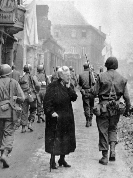Fassungslos steht eine alte Frau in den Trümmern der Stadt Bensheim, während US-Soldaten an ihr vorbei marschieren.