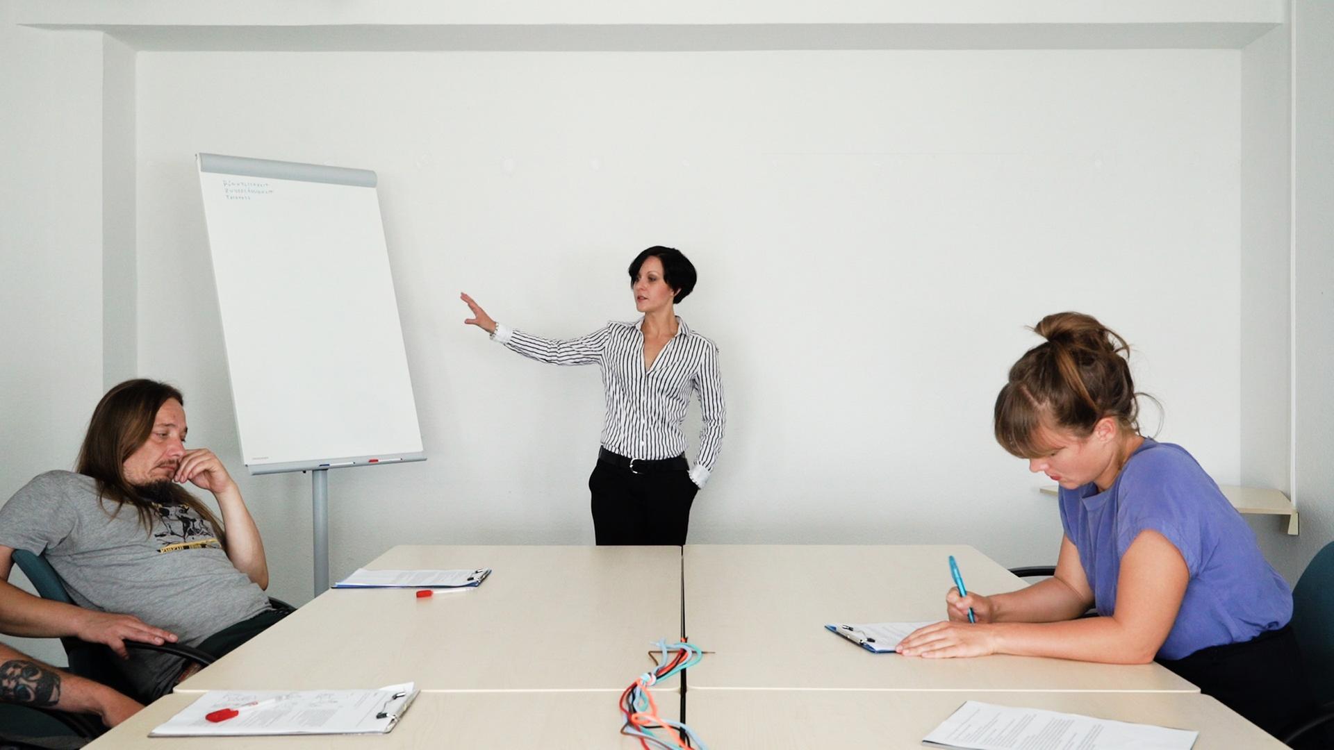 Ein Ausschnitt aus der Projektion "40h, max 2 Monate (work in progress)" von Stefanie Schröder. Eine Frau zeigt auf ein Flipchart, gelangweilte Teilnehmer der Maßnahme sitzen am Tisch Ein Ausschnitt aus der Projektion "40h, max 2 Monate (work in progress)" von Stefanie Schröder. Eine Frau zeigt auf ein Flipchart, gelangweilte Teilnehmer der Maßnahme sitzen am Tisch