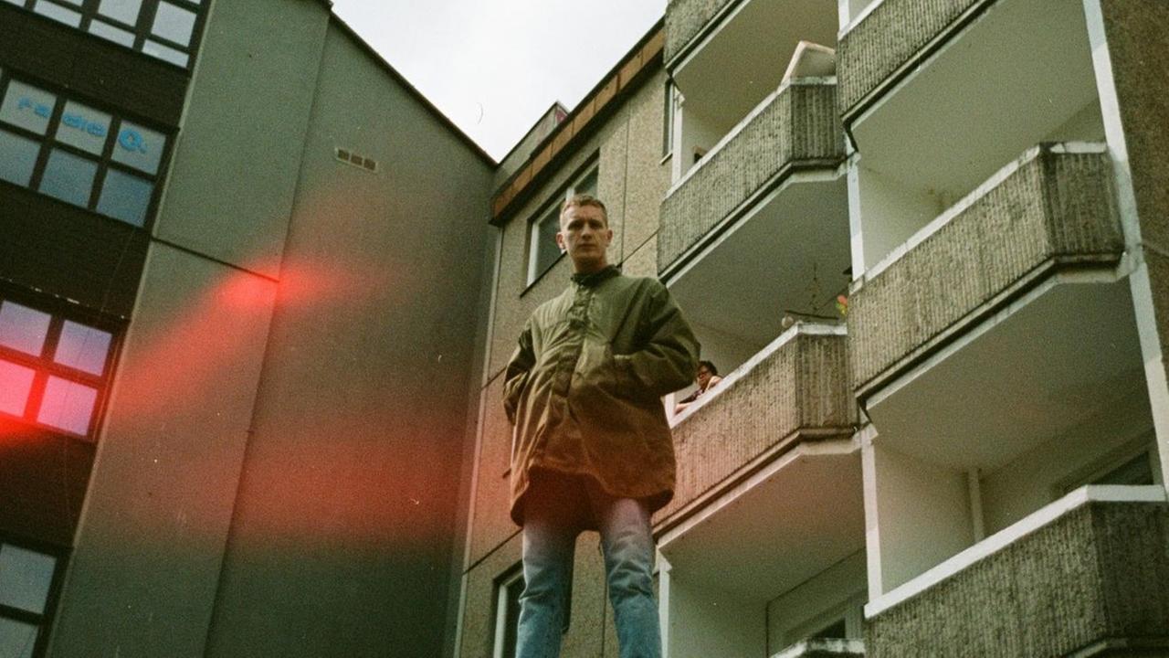Felix Kummer steht in einer grünen Jacke mit den Händen in den Taschen vor einem grünlich-grauen Plattenbau mit Balkonen. Felix Kummer steht in einer grünen Jacke mit den Händen in den Taschen vor einem grünlich-grauen Plattenbau mit Balkonen.