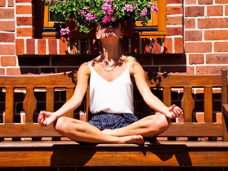 Eine junge Frau sitzt im Schneidersitzt auf einer Bank. Sieht atmet dabei den Geruch der Blumen über ihren Kopf tief ein.