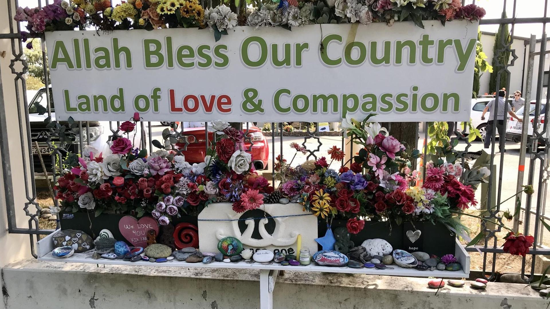 Vor einem Metallgitter liegen auf einer kleinen Mauer viele bemalte bunte Steine am Fuß von vielen Blumen und großen Blumensträußen. Darüber ein Plakat auf dem steht: "Allah schütze unser Land. Das Land von Liebe und Mitgefühl."