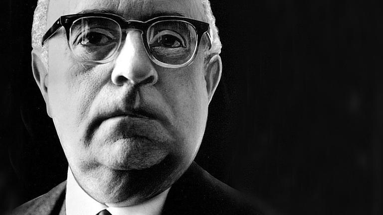 Ein Nahaufnahme von Theodor W. Adorno vor schwarzem Hintergrund. Er trägt eine Hornbrille und schaut aufmerksam dem Betrachter entgegen.