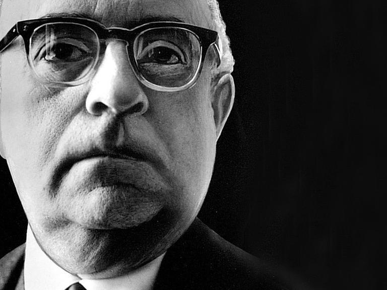Ein Nahaufnahme von Theodor W. Adorno vor schwarzem Hintergrund. Er trägt eine Hornbrille und schaut aufmerksam dem Betrachter entgegen.