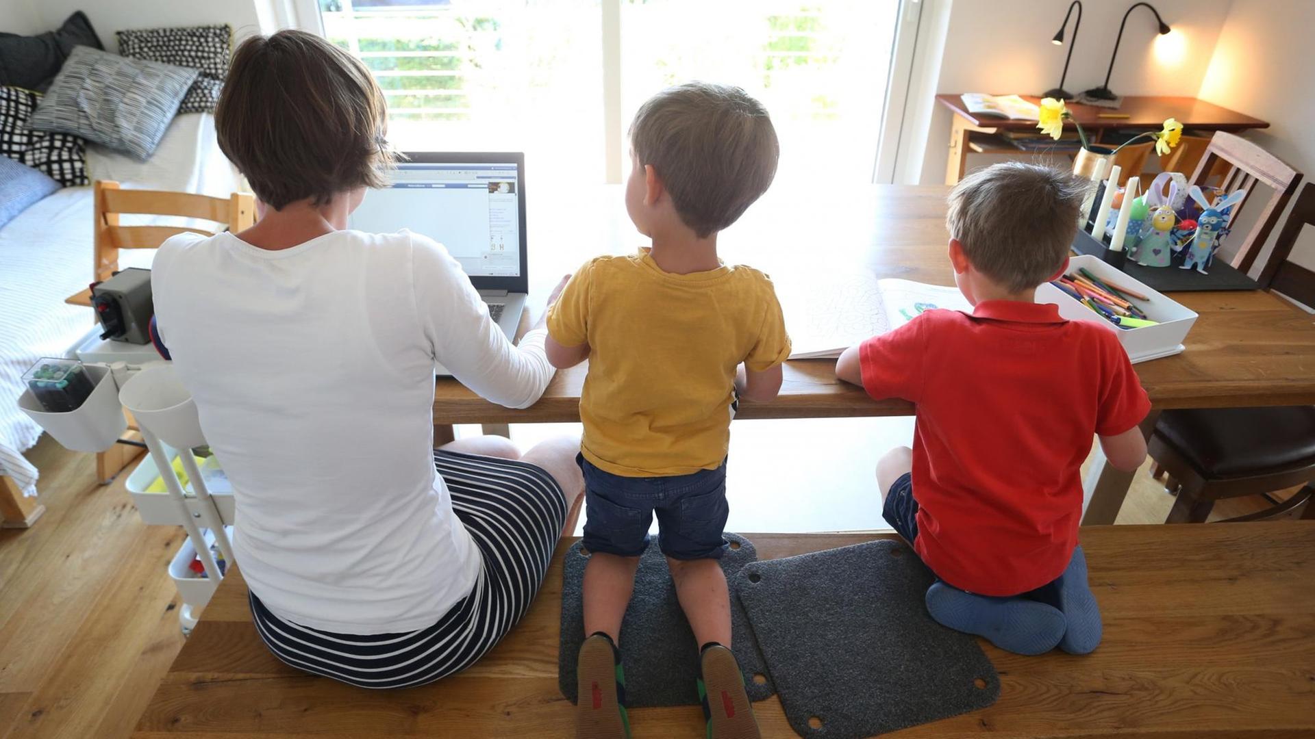 Eine Mutter sitzt mit ihren beiden kleinen Söhnen zuhause am Tisch, sie arbeitet am Laptop.