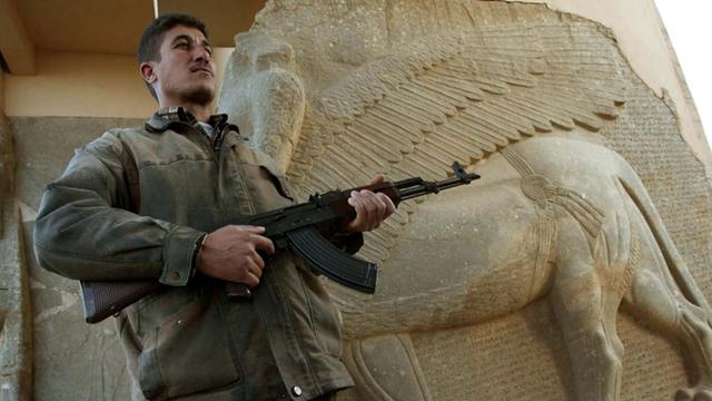 Ein mit einer Maschinenpistole bewaffneter Mann steht vor der steinernen Skulptur eines geflügelten Löwen.
