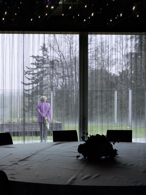 Doppelgängerin von Angela Merkel im Garten des Kanzlerbungalows. Im Vordergrund: Ein runder Konferenztisch mit Tischdecke.