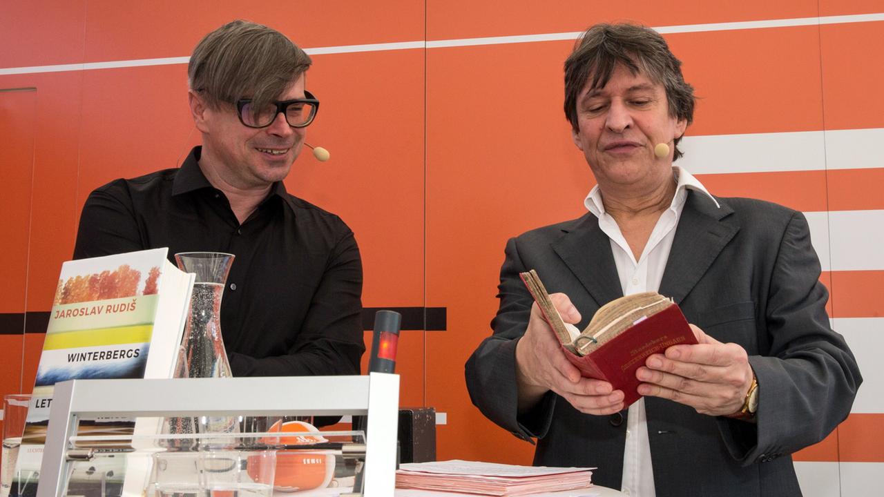 Autor Jaroslav Rudiš im Gespräch mit Moderator Joachim Scholl, der in einem alten Baedeker-Reiseführer blättert auf der Buchmesse in Leipzig 2019