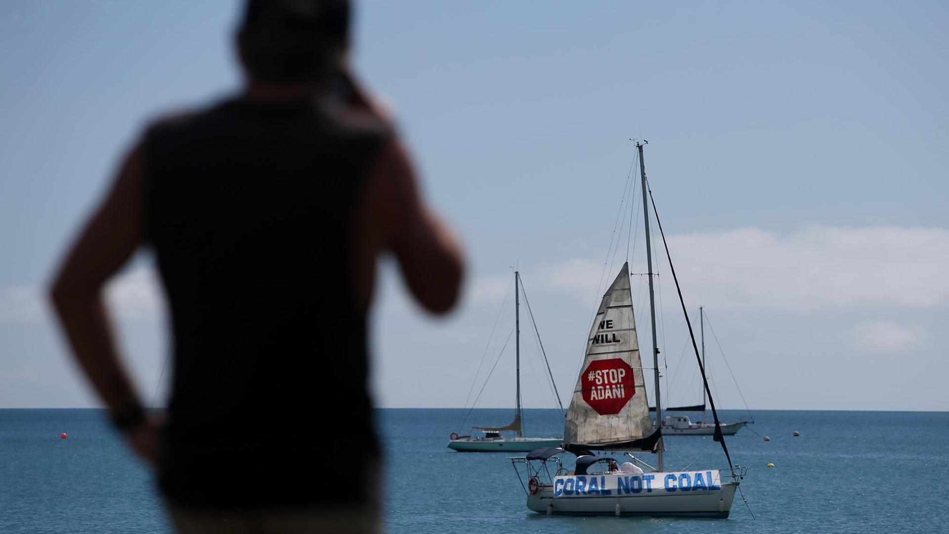 Protest gegen die Adani Kohlemine in Australien, Abbot Point: ein Segelschiff mit dem Banner "Stop Adani. Coral not Coal" liegt vor der Küste. Ein Mann im Vordergrund schaut auf das Meer, April 2019. Protest gegen die Adani Kohlemine in Australien, Abbot Point: ein Segelschiff mit dem Banner "Stop Adani. Coral not Coal" liegt vor der Küste. Ein Mann im Vordergrund schaut auf das Meer, April 2019.