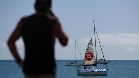 Protest gegen die Adani Kohlemine in Australien, Abbot Point: ein Segelschiff mit dem Banner "Stop Adani. Coral not Coal" liegt vor der Küste. Ein Mann im Vordergrund schaut auf das Meer, April 2019.