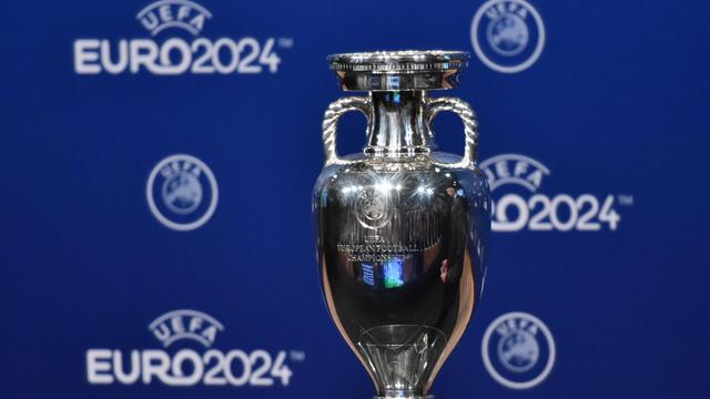 Das Foto zeigt den Pokal für die Fußball-Europameisterschaft, dahinter steht "Euro 2024" in weißer Schrift auf blauem Hintergrund.
