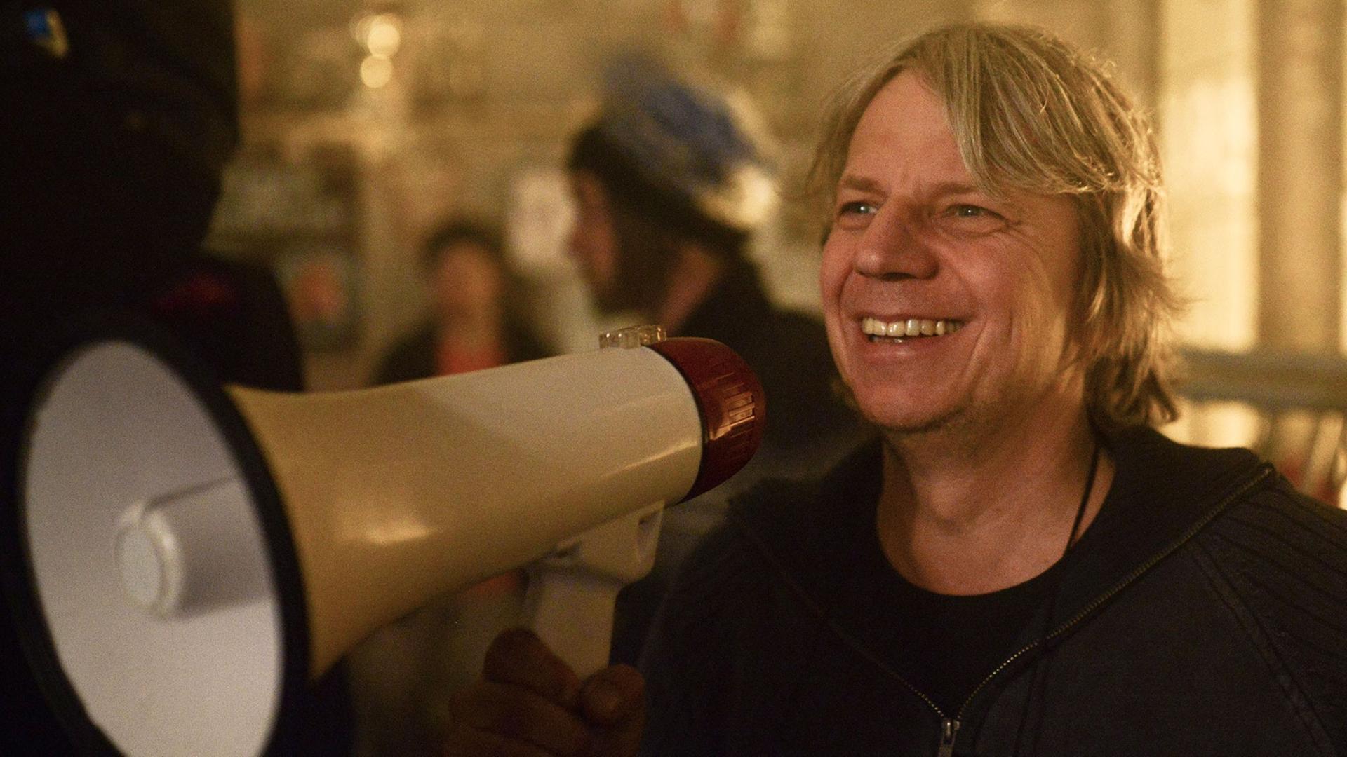 Regisseur Andreas Dresen hält lachend ein Megafon vor das Gesicht.