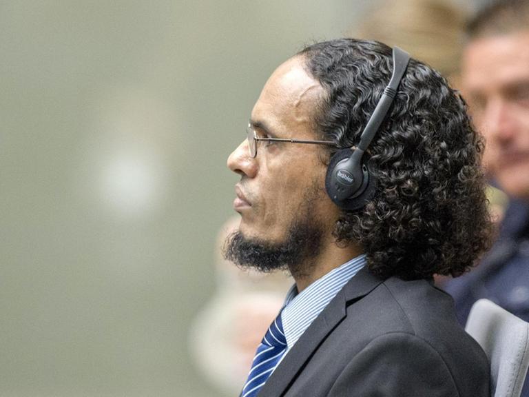 Das Bild zeigt Al Faqi Al Mahdi von der Seite auf der Anklagebank. Er trägt Kopfhörer und scheint jemandem aufmerksam zuzuhören. Er hat einen Anzug an und trägt eine Krawatte.