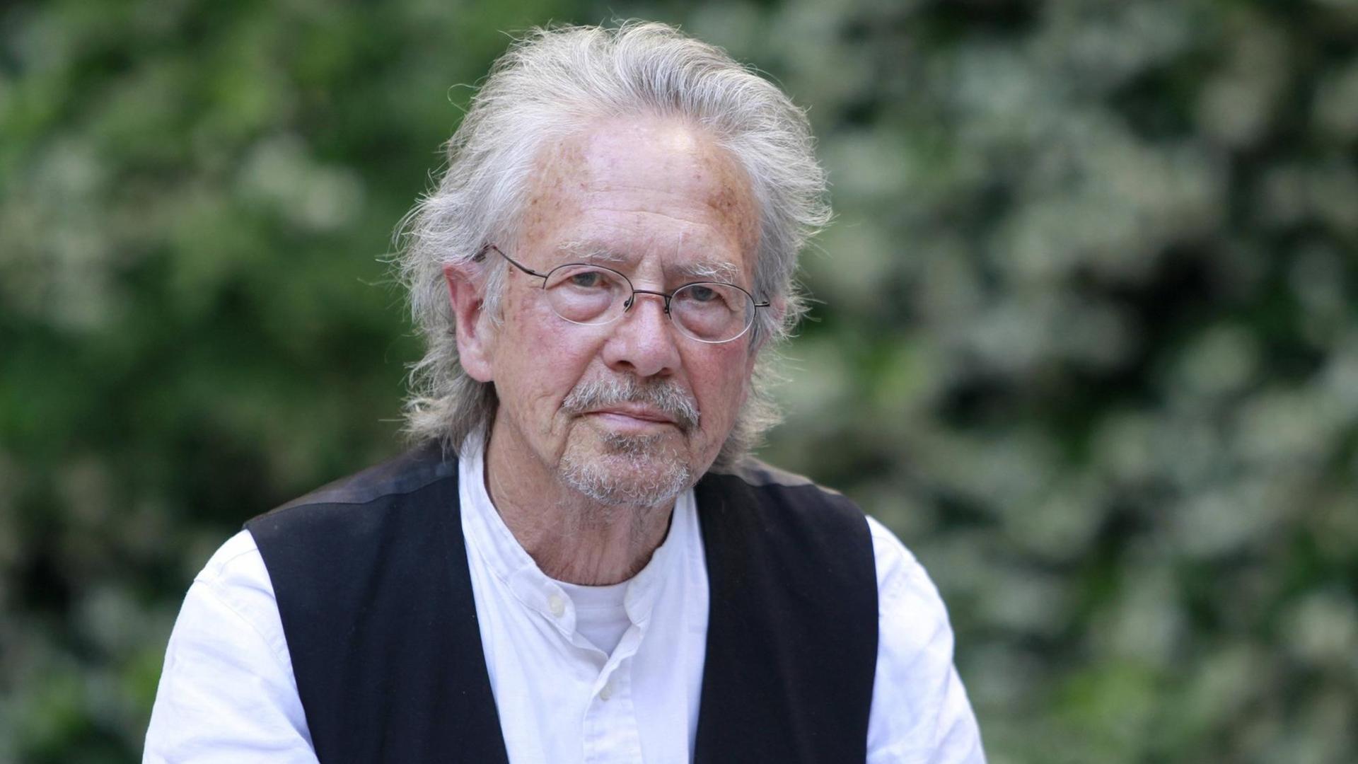 Peter Handke sitzt in weißem Hemd und schwarzer Wester vor einem unscharf gestellten Gebüsch und blickt freundlich in die Kamera Peter Handke sitzt in weißem Hemd und schwarzer Wester vor einem unscharf gestellten Gebüsch und blickt freundlich in die Kamera