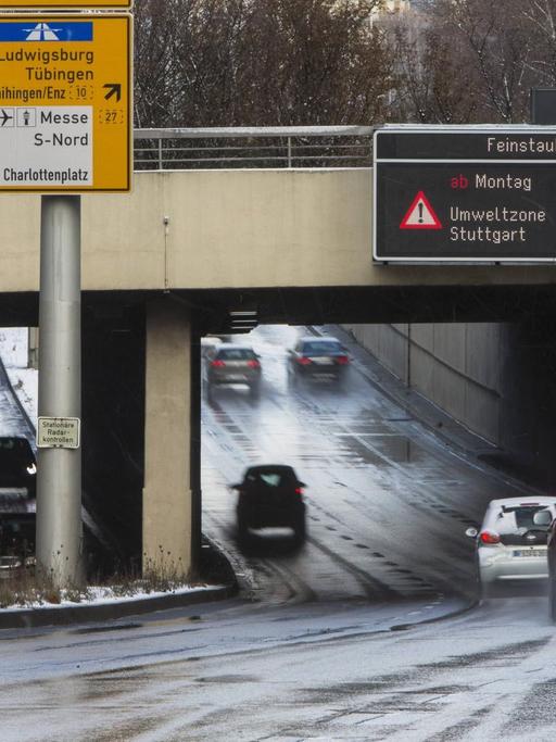 Autos fahren unter einer Brücke hindurch, an der ein Warnschild mit der Aufschrift "Feinstaub-Alarm Umweltzone Stuttgart - bitte Busse und Bahnen nutzen" hängt.