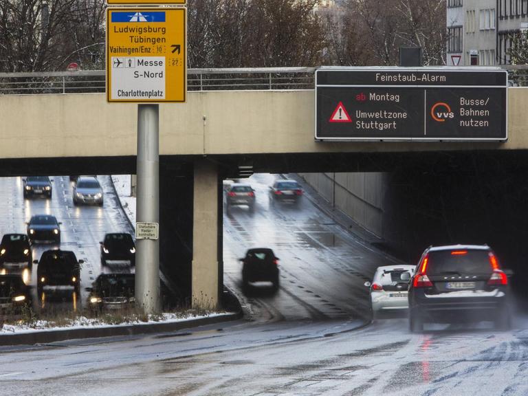 Autos fahren unter einer Brücke hindurch, an der ein Warnschild mit der Aufschrift "Feinstaub-Alarm Umweltzone Stuttgart - bitte Busse und Bahnen nutzen" hängt.