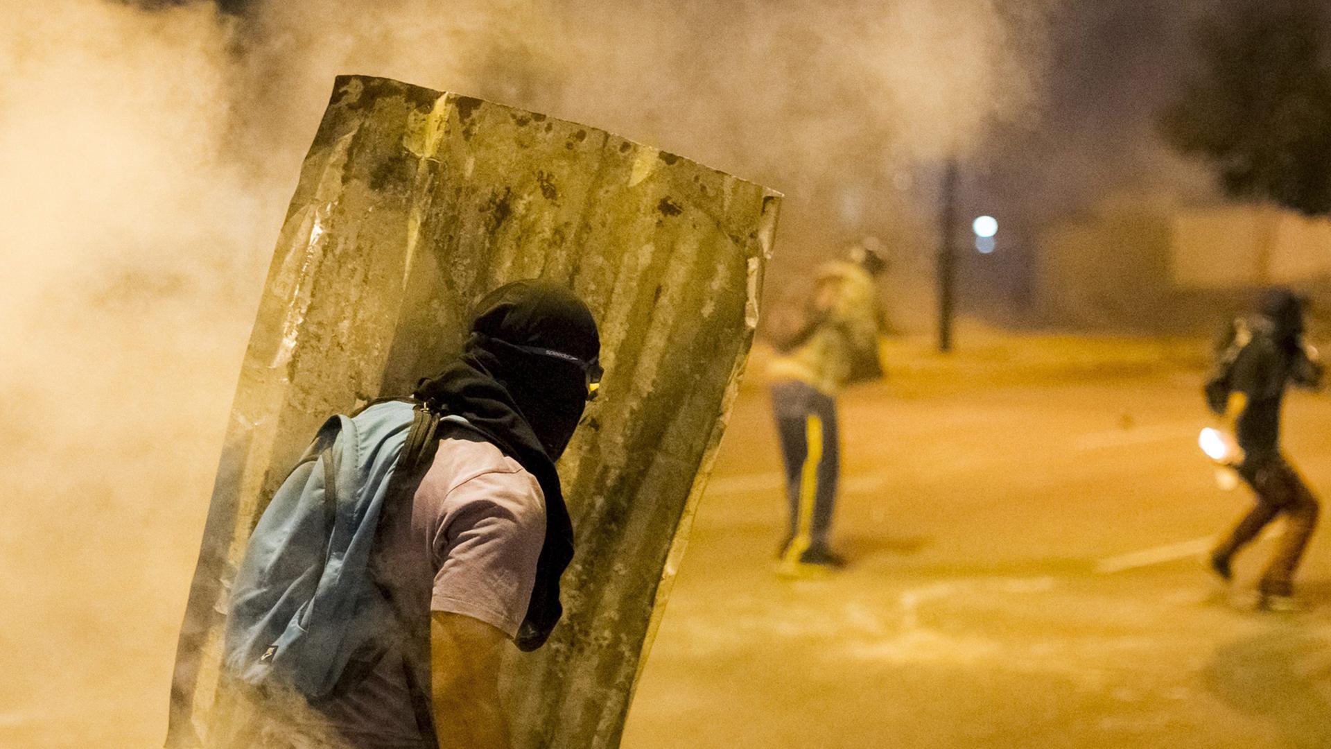 In der venezuelanischen Stadt Altamira ist am 24.02.2014 ein vermummter Demonstrant zu sehen, der einen Schild aus Wellblech trägt und von Qualm umgeben ist. In der venezuelanischen Stadt Altamira ist am 24.02.2014 ein vermummter Demonstrant zu sehen, der einen Schild aus Wellblech trägt und von Qualm umgeben ist.