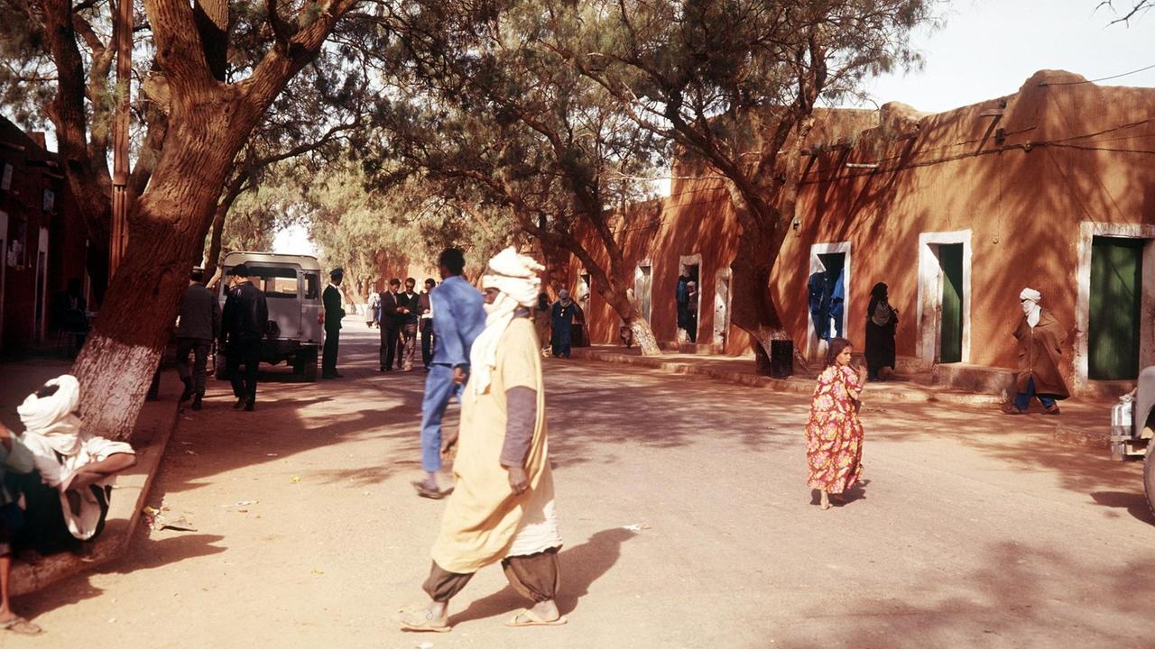 Blick in die Hauptstraße von Tamanrasset, dem Hauptort der Tuareg. Mehrere Menschen, u.a. mit Turbanen bekleidet, laufen über einen Platz mit einem großen Baum und einem Haus im Hintergrund. Blick in die Hauptstraße von Tamanrasset, dem Hauptort der Tuareg. Mehrere Menschen, u.a. mit Turbanen bekleidet, laufen über einen Platz mit einem großen Baum und einem Haus im Hintergrund.