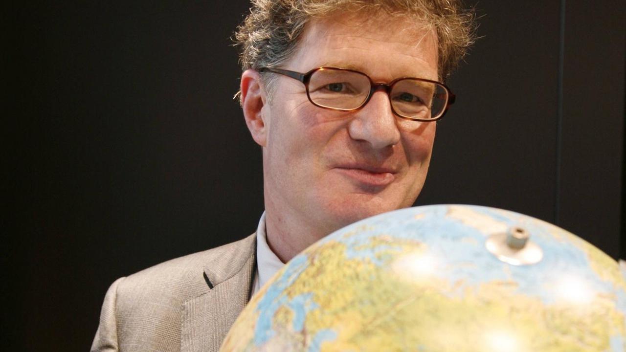 Ein Mann mit Brille hält einen Globus vor die Brust und schaut lächelnd darüber hinweg. Ein Mann mit Brille hält einen Globus vor die Brust und schaut lächelnd darüber hinweg.