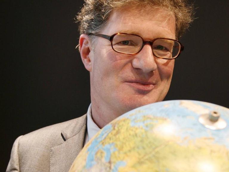Ein Mann mit Brille hält einen Globus vor die Brust und schaut lächelnd darüber hinweg.