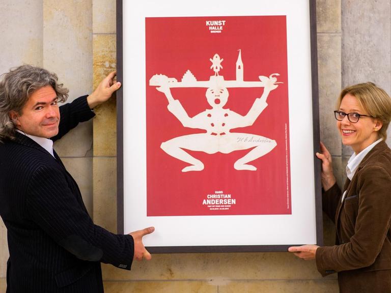 Anne Buschhoff und Detlef Stein stehen in der Bremer Kunsthalle und halten das eingerahmte Scherenschnittplakat von Hans Christian Andersen an eine Wand