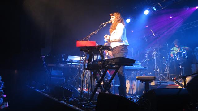 Eine Frau mit langen schwarzen Haaren steht hinter einem E-Piano auf einer Konzertbühne und spricht zu den Zuschauern.