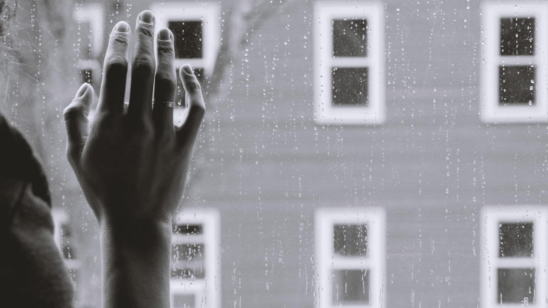 Schwarz-weiß-Bild einer Frauenhand an einer Fensterscheibe, draußen regnet es. Die Frau, deren Gesicht man nicht sieht, schaut offenbar auf die Straße. Schwarz-weiß-Bild einer Frauenhand an einer Fensterscheibe, draußen regnet es. Die Frau, deren Gesicht man nicht sieht, schaut offenbar auf die Straße.