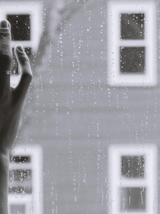 Schwarz-weiß-Bild einer Frauenhand an einer Fensterscheibe, draußen regnet es. Die Frau, deren Gesicht man nicht sieht, schaut offenbar auf die Straße.