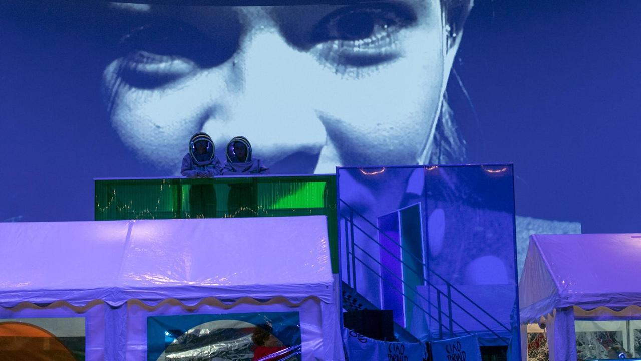 Auf einer Theaterbühne stehen im Vordergrund zwei blaue Plastikzelte. Dahinter stehen auf einer erhöhten Treppe zwei Darsteller in Weltraumanzügen und mit Astronautenhelmen, die sich dem Publikum zuwenden. Auf der Wand dahinter ist in Nahaufnahme das Gesicht der Schauspielerin Paula Kober projiziert. Auf einer Theaterbühne stehen im Vordergrund zwei blaue Plastikzelte. Dahinter stehen auf einer erhöhten Treppe zwei Darsteller in Weltraumanzügen und mit Astronautenhelmen, die sich dem Publikum zuwenden. Auf der Wand dahinter ist in Nahaufnahme das Gesicht der Schauspielerin Paula Kober projiziert.