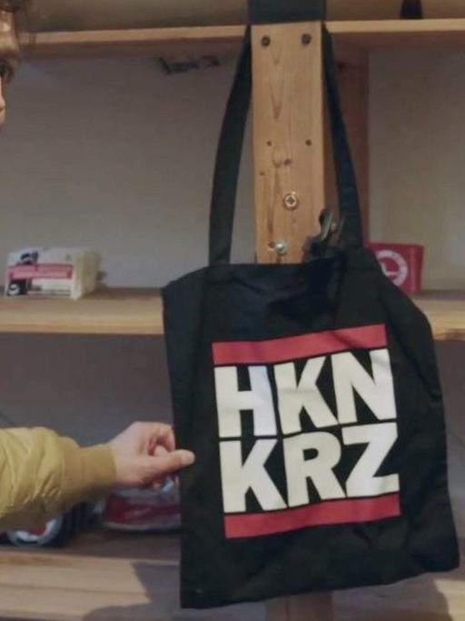 Pro7-Reporter Thilo Mischke steht vor einem Regal und zeigt eine schwarze Stofftasche, die mit der Abkürzung "HKN KRZ" für "Hakenkreuz" bedruckt ist.