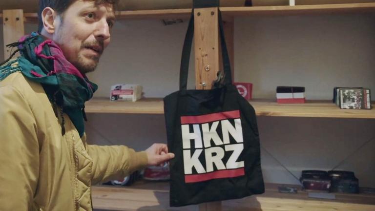 Pro7-Reporter Thilo Mischke steht vor einem Regal und zeigt eine schwarze Stofftasche, die mit der Abkürzung "HKN KRZ" für "Hakenkreuz" bedruckt ist.