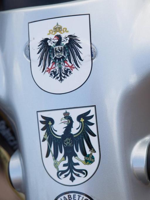 Mehrere Wappen als Statement für eine Monarchie nach Vorbild des Deutschen Reichs kleben auf einem kleinen Wagen für Menschen mit Gehproblemen.