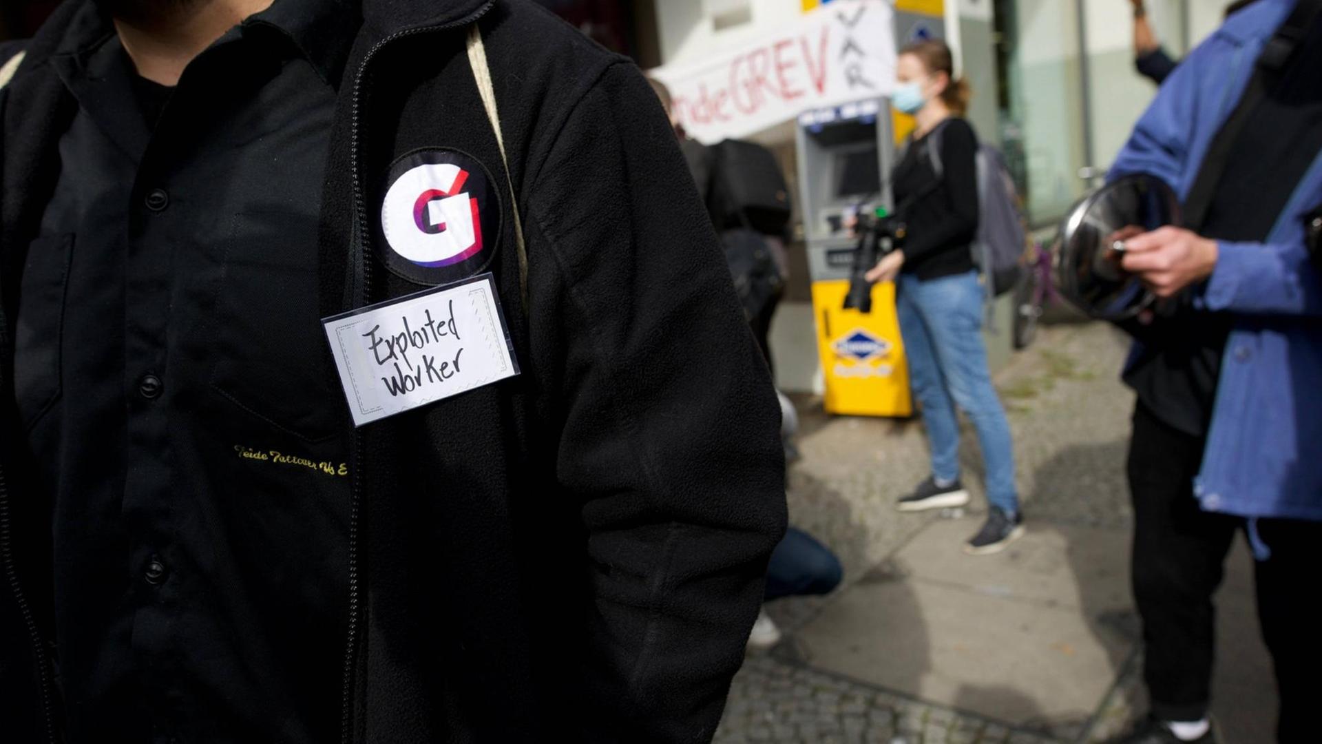 Im Vordergrund eine Person mit schwarzer Jacke, die das „G“ der Firma Gorillas als Sticker auf der Jacke trägt. Darunter ein Klebestreifen mit der Aufschrift „Exploited Worker“ Im Vordergrund eine Person mit schwarzer Jacke, die das „G“ der Firma Gorillas als Sticker auf der Jacke trägt. Darunter ein Klebestreifen mit der Aufschrift „Exploited Worker“