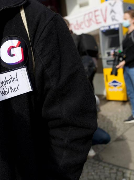 Im Vordergrund eine Person mit schwarzer Jacke, die das „G“ der Firma Gorillas als Sticker auf der Jacke trägt. Darunter ein Klebestreifen mit der Aufschrift „Exploited Worker“