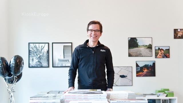 Markus Schaden, Fotograf und Kurator, dunkle Haare, dunkles Hemd, Brille, steht lächelnd hinter einem Tisch mit Fotobüchern, an der weißen Wand hinter ihm hängen Bilder