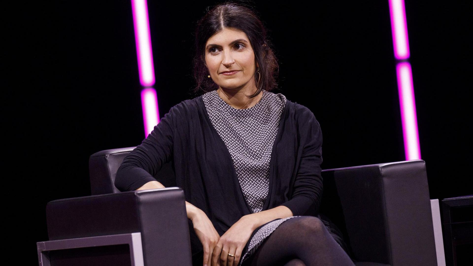 Shida Bazyar auf einem Sessel auf einer Bühne, im Hintergrund sind rosa Neonröhren zu sehen Shida Bazyar auf einem Sessel auf einer Bühne, im Hintergrund sind rosa Neonröhren zu sehen
